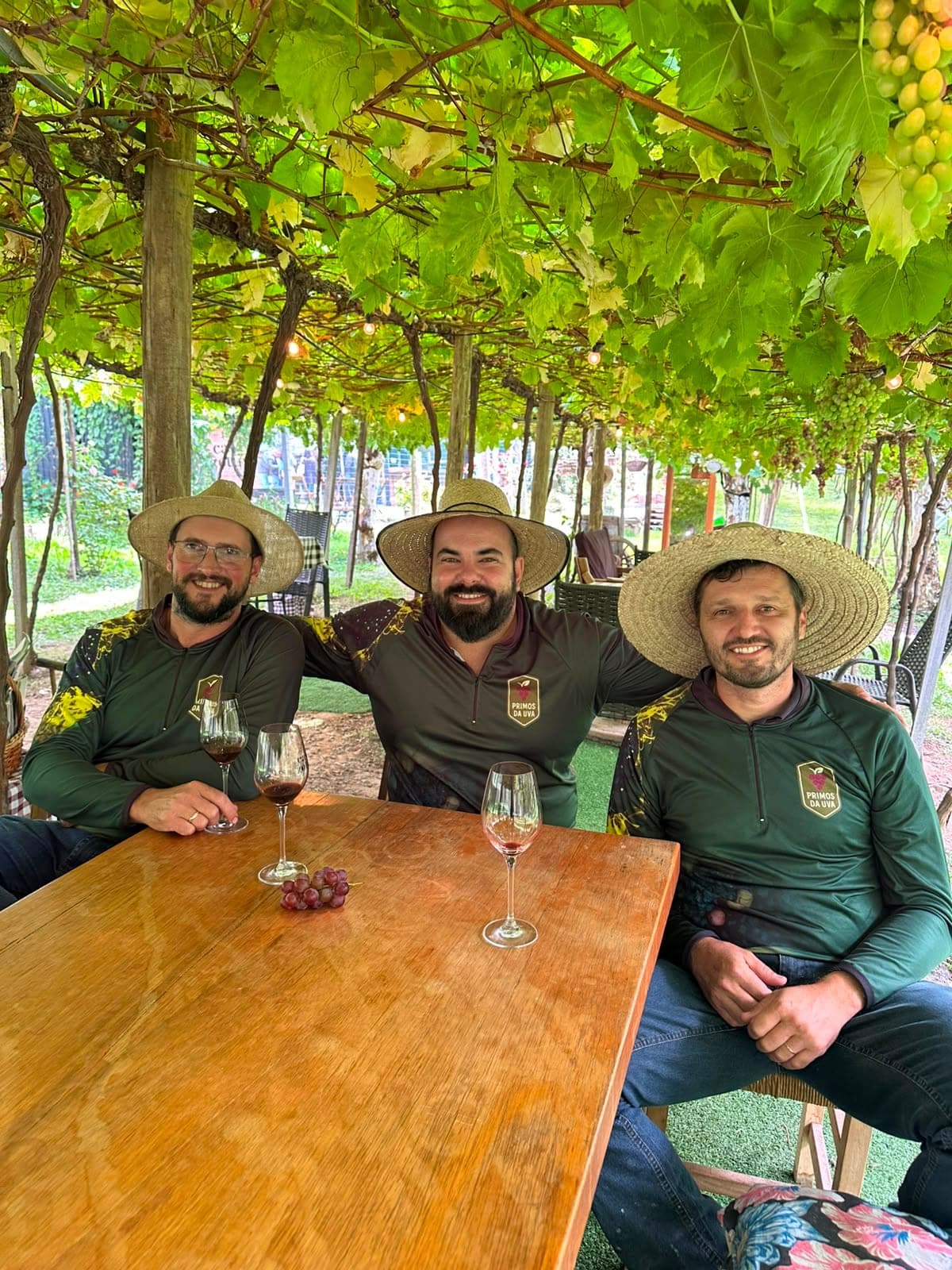 Sócios da Primos da Uva reunidos sob o parreiral, de chapéu de palha e camisa da marca